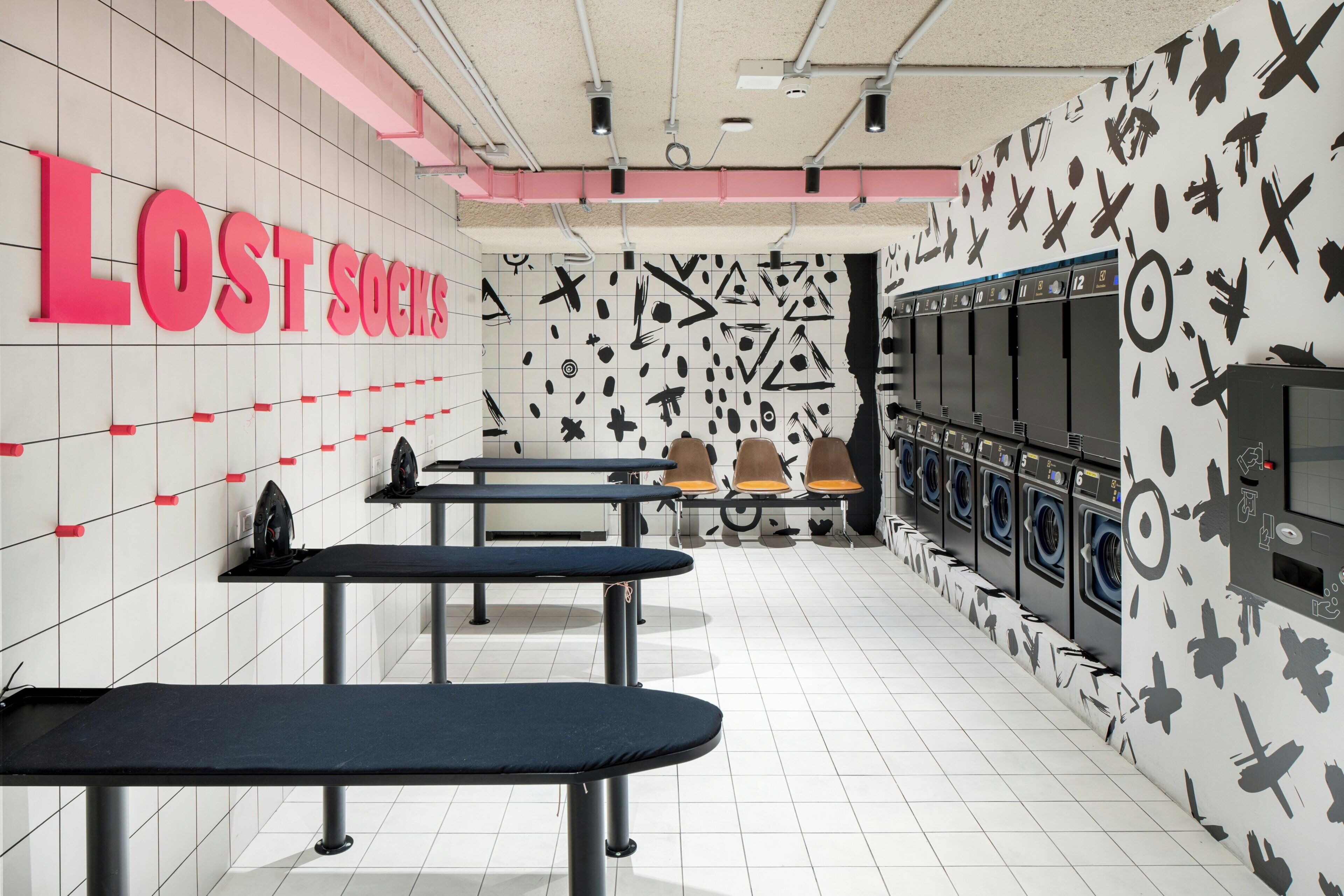 A laundromat with rows of washing machines, ironing stations, and a wall with a geometric pattern.