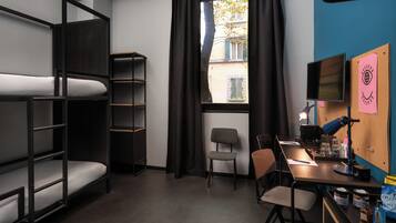 A dorm-style room with bunk beds, a desk, a chair, a computer monitor, and a window with curtains.