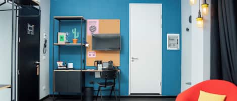 A compact room with a desk, chair, and a red chair with a cushion. There is a door, a shelf with items, and a vent on the wall.