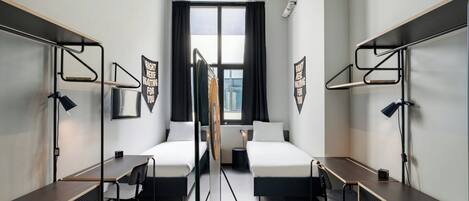 A modern hotel room with two single beds, a desk, chairs, a window with curtains, and a view of the city.