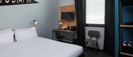 A hotel room with a bed, a desk with a computer, a chair, a TV, and a window with curtains.