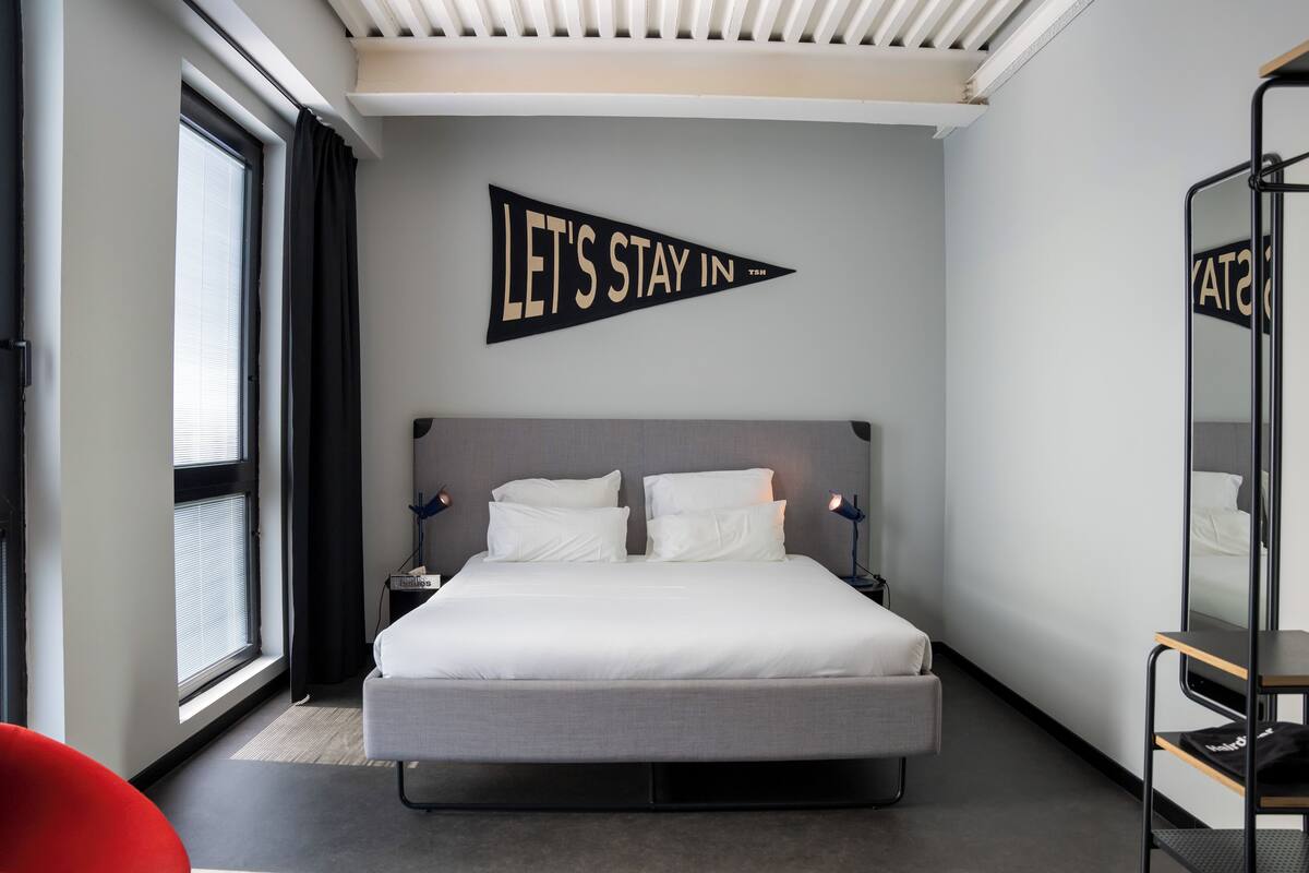 Une chambre d’hôtel moderne avec un grand lit, une table de chevet, un miroir et une fenêtre avec des rideaux.