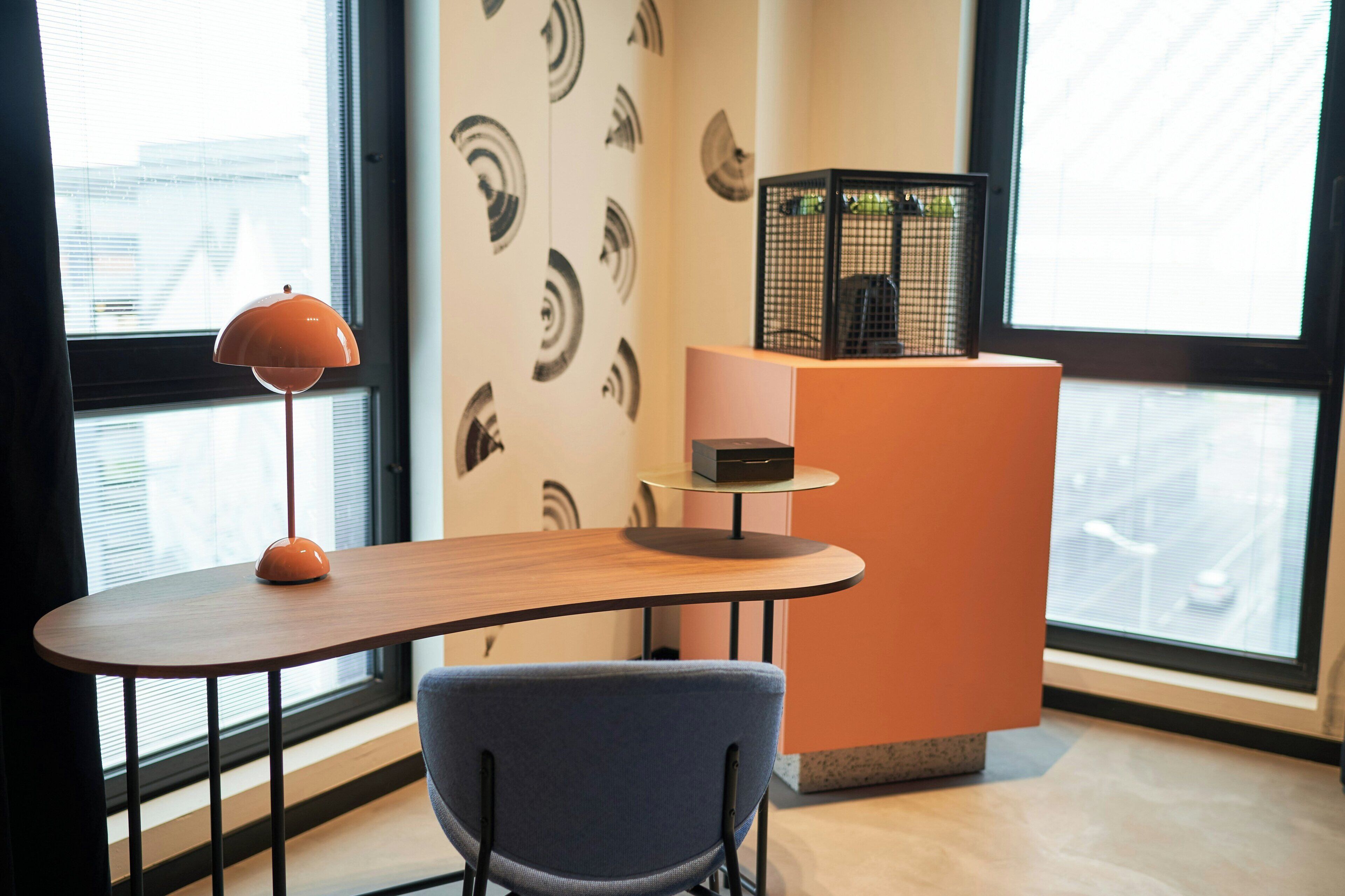 A modern room with a curved wooden table, a blue chair, a floor lamp, and a decorative black cage on a pedestal.