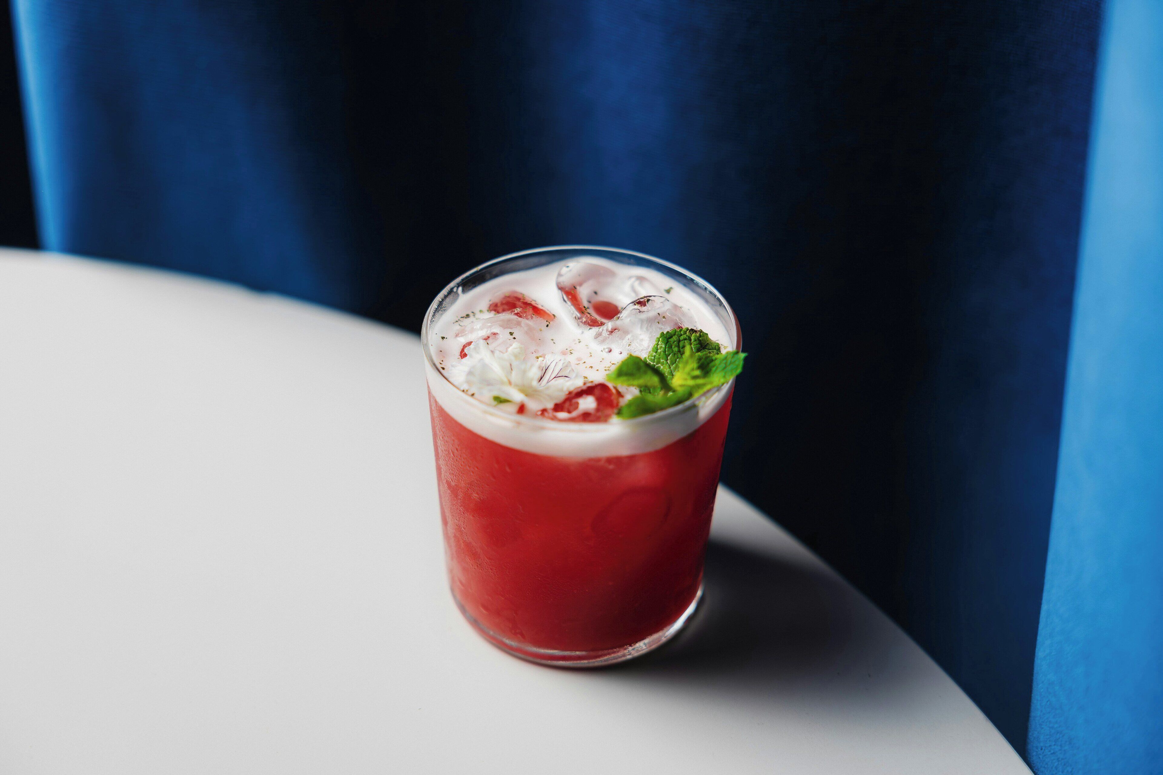 A glass of red beverage with ice and a mint garnish on a white surface.