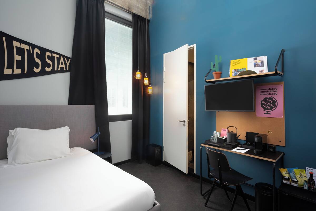 Une chambre d’hôtel avec un lit, un bureau, une chaise et un mur sur lequel est affiché un panneau indiquant : « RESTEZ ENCORE UN PEU ».