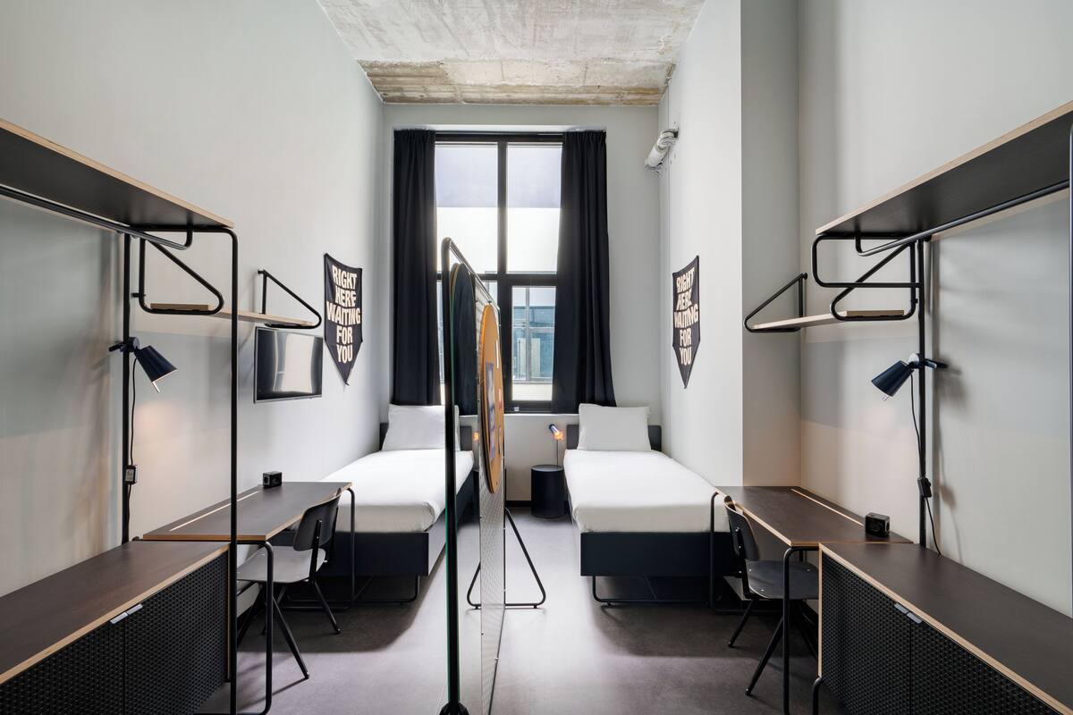 Une chambre d’hôtel moderne avec deux lits simples, un bureau et une chaise. Il y a deux lampes noires, une de chaque côté de la pièce.