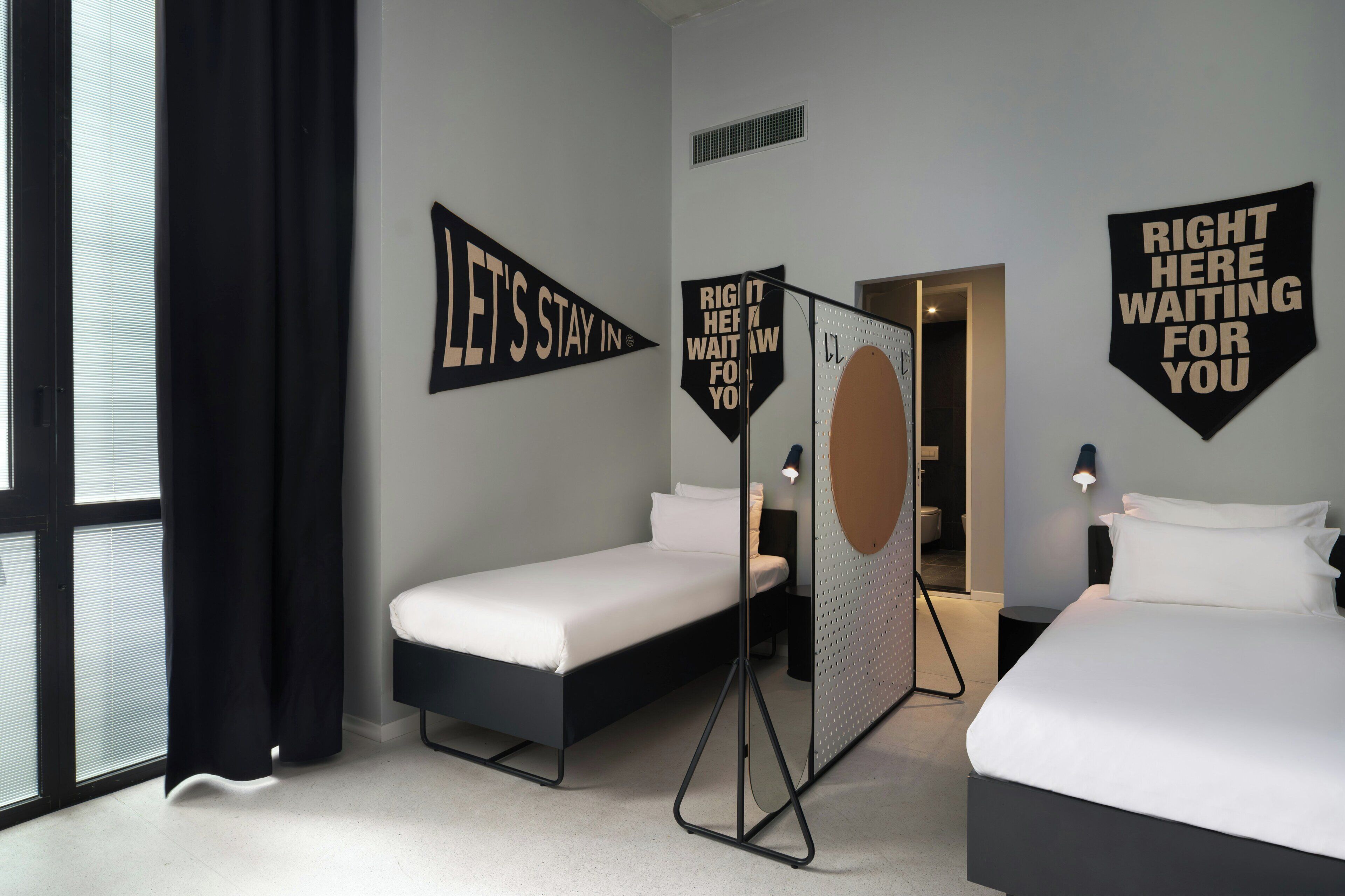 Une chambre d’hôtel avec deux lits, une grande fenêtre avec des rideaux noirs et des œuvres d’art motivantes accrochées aux murs.