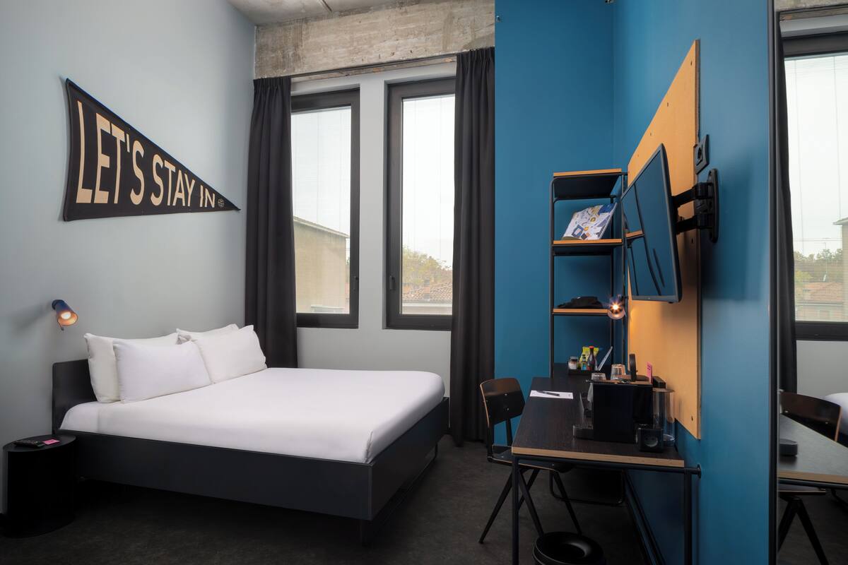 Une chambre d’hôtel moderne avec un grand lit, un bureau avec une chaise, un téléviseur fixé au mur et une pancarte indiquant « Restons à l’hôtel ».