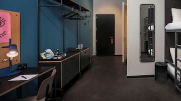 Quarto de hotel compacto com escrivaninha, cadeira, beliches, espelho e cabideiro.