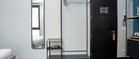 A compact room with a bed, a mirror, a black wardrobe, a small shelf, and a heater.
