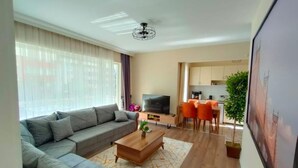 A modern living room with a sofa, coffee table, and TV. There is a dining area with chairs and a table. - Lara Park Suites (Antalya)