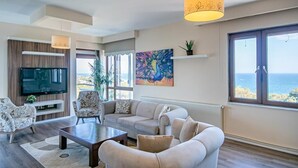 A modern living room with a large TV, comfortable seating, a painting, and a view of the sea. - Lara Park Suites (Antalya)