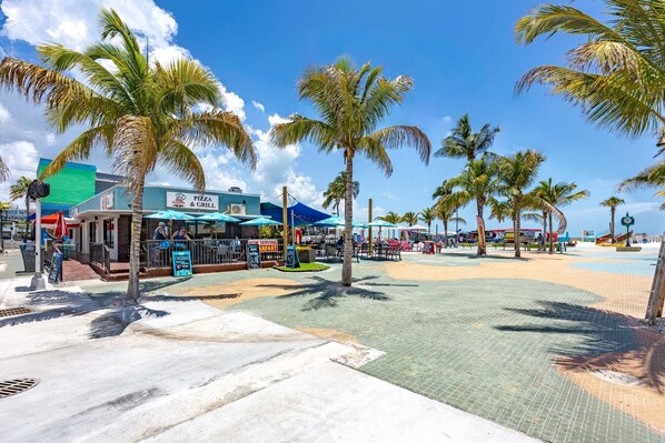 Zona frente a la playa con restaurante de pizza y parrilla, palmeras y un paseo de baldosas.
