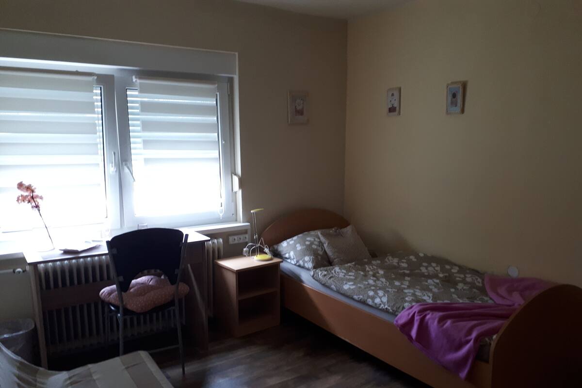 A small, well-lit room with a single bed, a chair, a desk, and a radiator.