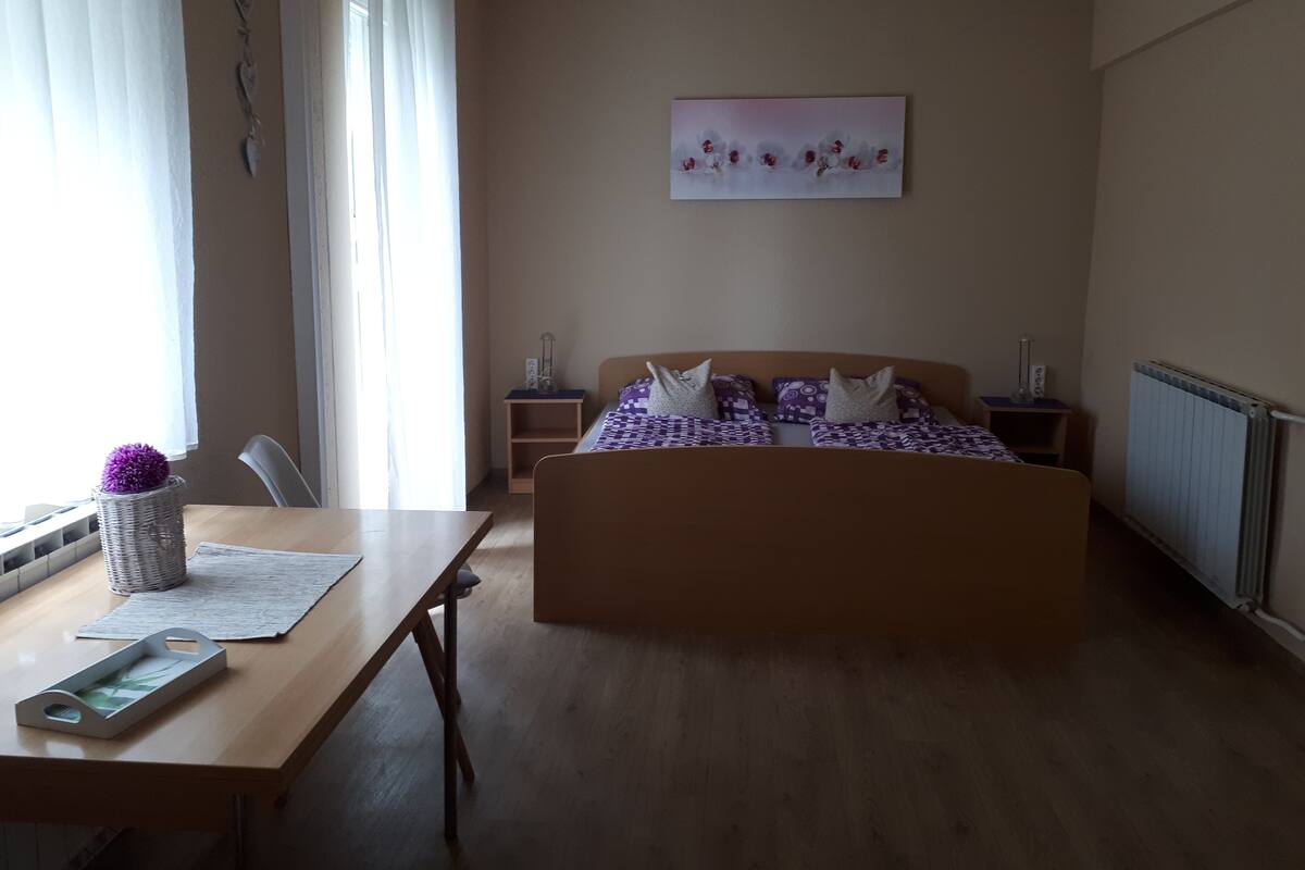 A bedroom with a wooden bed, a bedside table, a chair, a window with curtains, and a wall-mounted artwork.