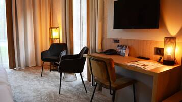 A hotel room with a bed, two chairs, a desk with a lamp, a TV, and a window with curtains.