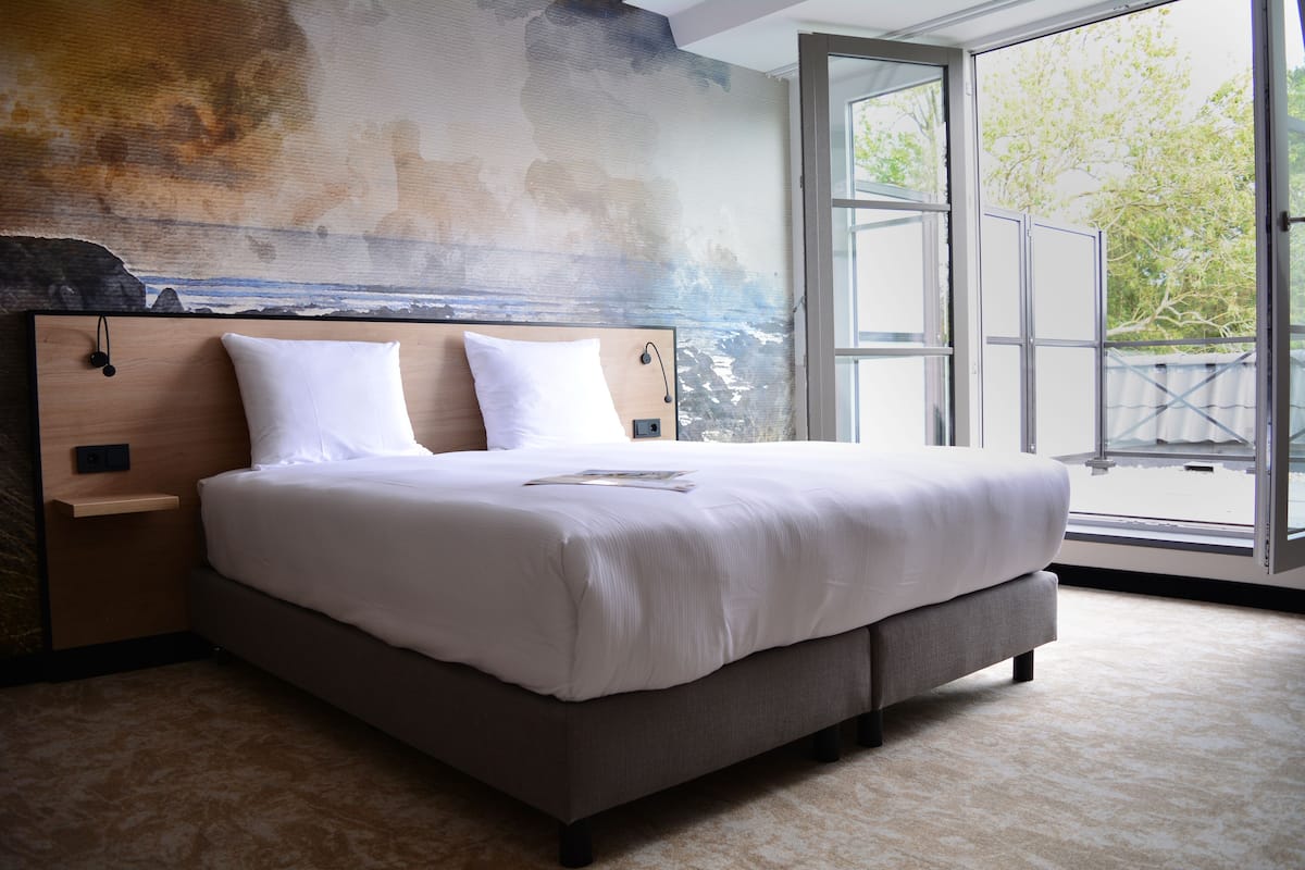 A modern bedroom with a large bed, a wooden headboard, and a view of the outdoors through a large window.