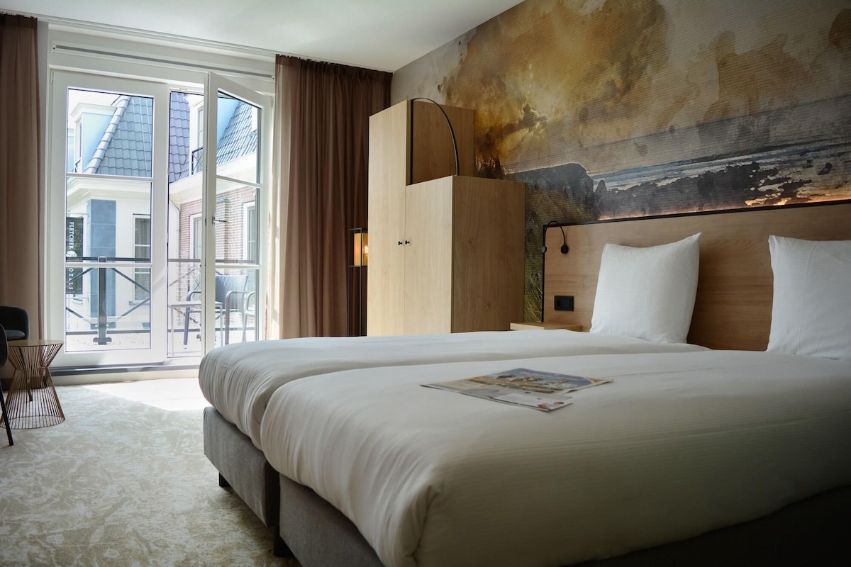 A hotel room with a large bed, a view of the city through a glass door, and a painting on the wall.