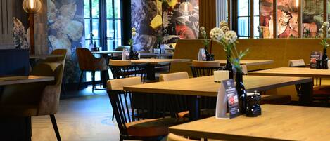 A restaurant with wooden tables and chairs, large windows, and a mural on the wall.