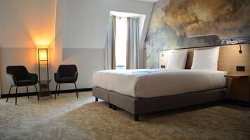 A modern hotel room with a large bed, two chairs, a small table, and a large abstract painting on the wall.
