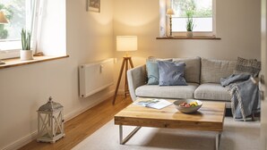 Un salon chaleureux avec une table basse en bois, un canapé gris agrémenté de coussins, une lampe sur pied et une fenêtre ornée d’une plante.