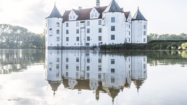 Un grand château blanc, doté de nombreuses tours, qui se reflète dans l’eau calme.