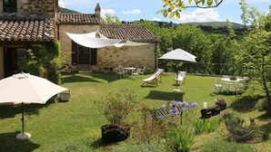 Un edificio de piedra con tejado de baldosas, mesas al aire libre con sombrillas, y un jardín con césped y flores.
