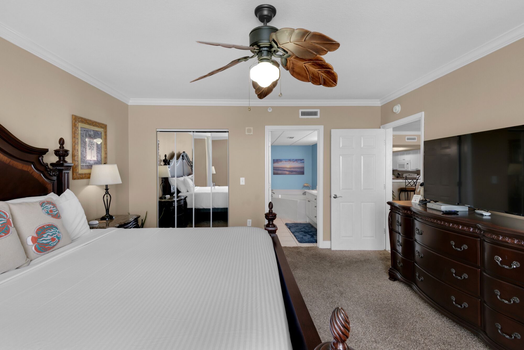 Une chambre d’hôtel avec un grand lit, un ventilateur de plafond, une commode, un téléviseur et une salle de bain visible à travers une porte ouverte.
