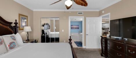 Une chambre d’hôtel avec un grand lit, un ventilateur de plafond, une commode, un téléviseur et une salle de bain visible à travers une porte ouverte.