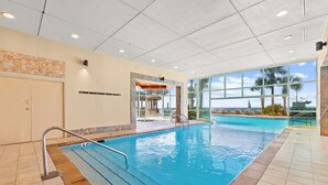 Una piscina interior con paredes de vidrio transparente, suelo de baldosas y vistas a palmeras y al océano.
