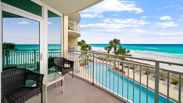 Un balcon avec vue sur une piscine et une plage, des fauteuils en osier et une table en verre.