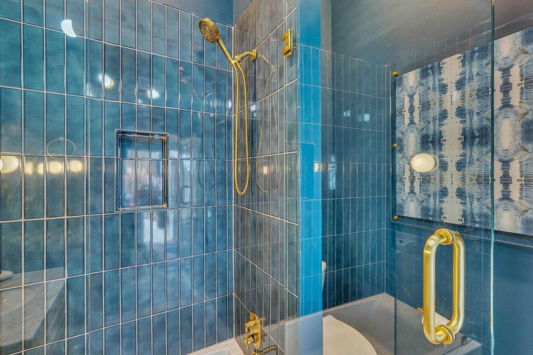 Un baño moderno con paredes de azulejos azules, zona de ducha con mampara de cristal y cabezal de ducha dorado.
