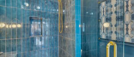 Un baño moderno con paredes de azulejos azules, zona de ducha con mampara de cristal y cabezal de ducha dorado.