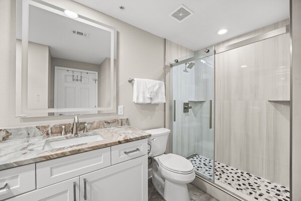 Un baño moderno con encimera de mármol, inodoro, cabina de ducha de vidrio y un espejo grande.