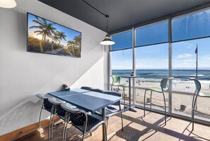 Quarto com uma grande janela com vista para a praia, uma mesa com cadeiras e um quadro na parede.