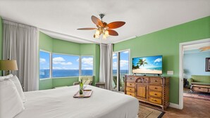 A hotel room with a large bed, a ceiling fan, a television, and a view of the ocean.