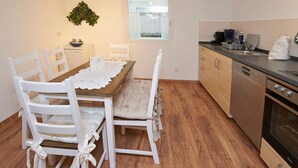 A compact kitchen with wooden cabinets, a dishwasher, and a stove. There is a dining table with white chairs and a lace tablecloth. A window with a view of a building is visible. - Spacious apartment in the old town, 90 sqm, 1st sep. Bedroom (Wasserburg am Inn)
