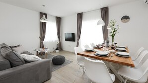 Una sala moderna con un sofá gris, una mesa de comedor de madera preparada para seis personas, sillas blancas, un televisor montado en la pared y un amplio ventanal con cortinas.