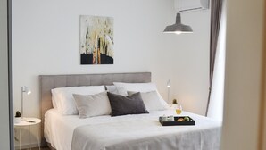 Un dormitorio moderno con una cama grande, una mesita de noche con lámpara, una pequeña mesa con una planta y un cuadro en la pared.