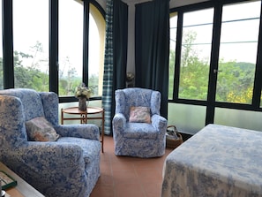 A room with two patterned armchairs, a small round table with a vase, and a view of greenery through large windows.