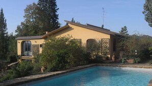 Una piscina con baldosas de piedra, rodeada de vegetación y un edificio con fachada amarilla y un balcón.