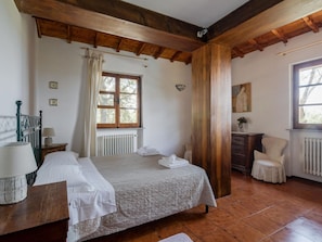 Een slaapkamer met een houten plafond, een bed met quilt, een nachtkastje met lamp, een stoel en een raam met gordijnen.
