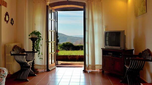 A room with a large open doorway leading to a scenic outdoor view, a television on a wooden cabinet, and a potted plant near the doorway. - Elegant Villa Near Cortona (Cortona)