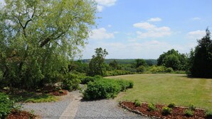 A pathway leading through a landscaped garden with a variety of plants and trees, a clear sky, and a distant view of a hilly landscape. - Holiday Home in Noiseux With Private Hot Tub (Somme-Leuze)