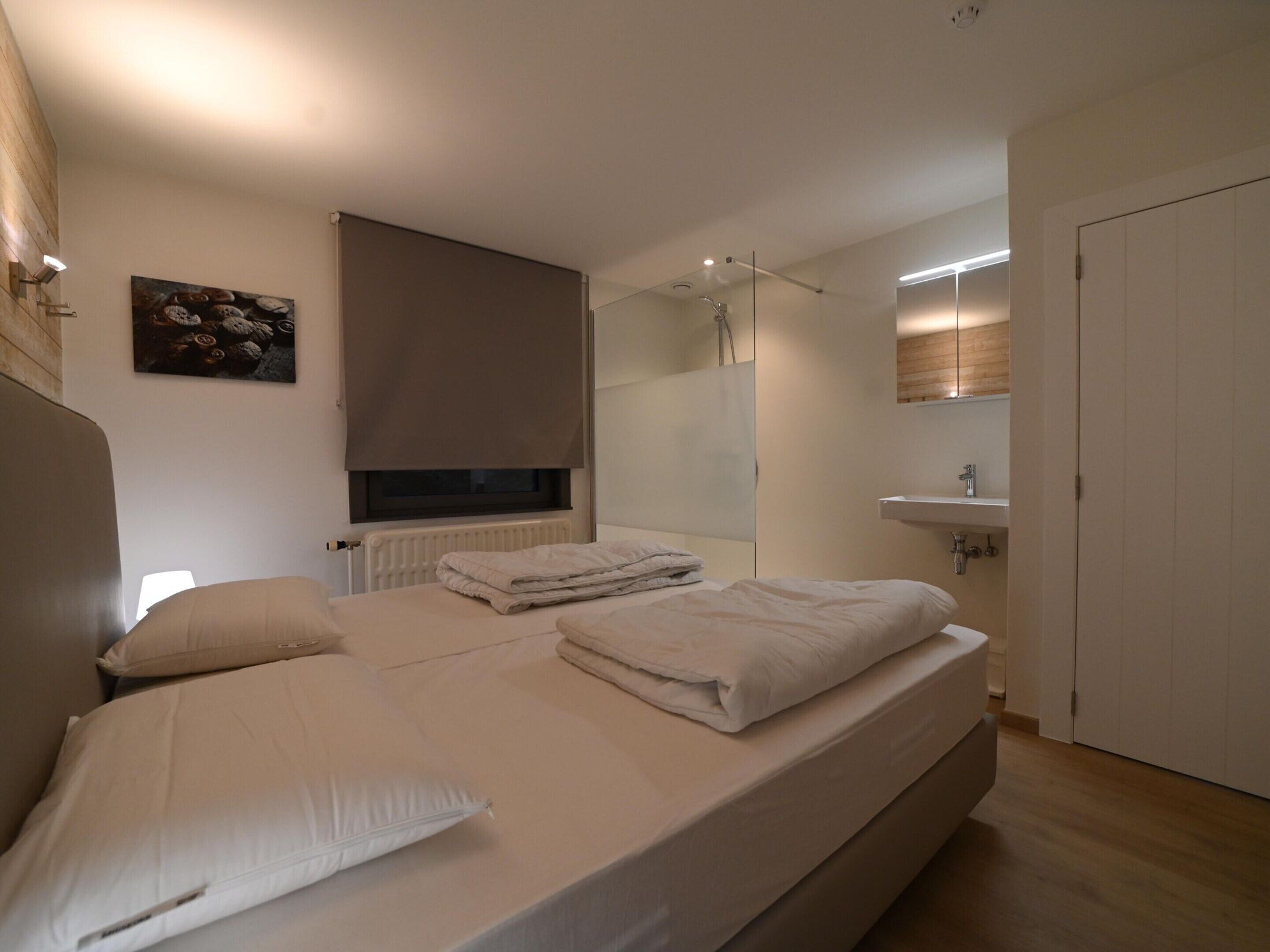 A bedroom with a bed, a window with blinds, a radiator, a mirror, a sink, and a door.