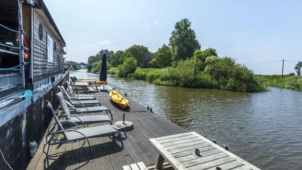 A wooden deck with lounge chairs and a kayak, overlooking a calm river with greenery on the banks.
