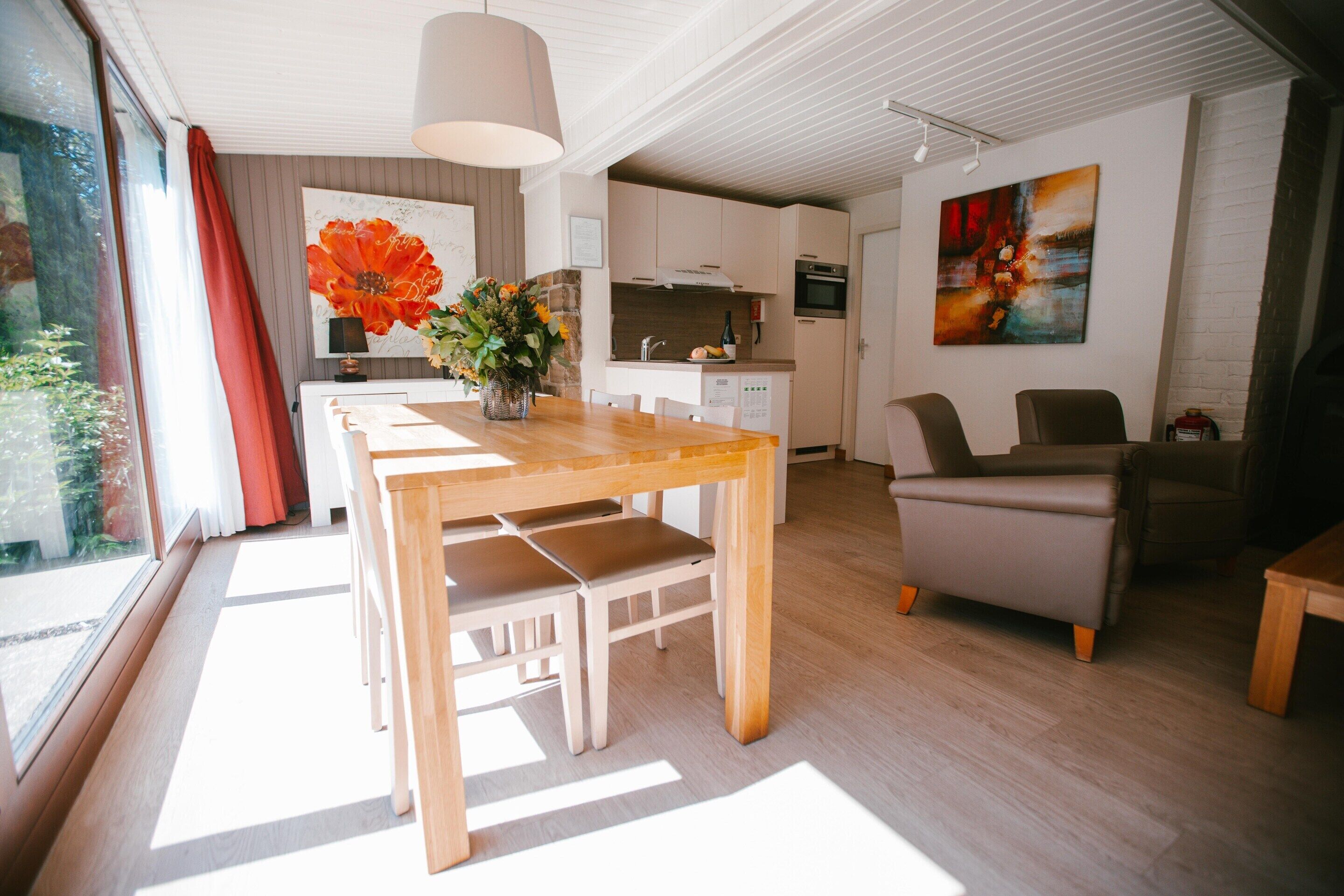Een moderne keuken met witte kasten, een eethoek met een houten tafel en stoelen, en een woonkamer met een bank en fauteuils.
