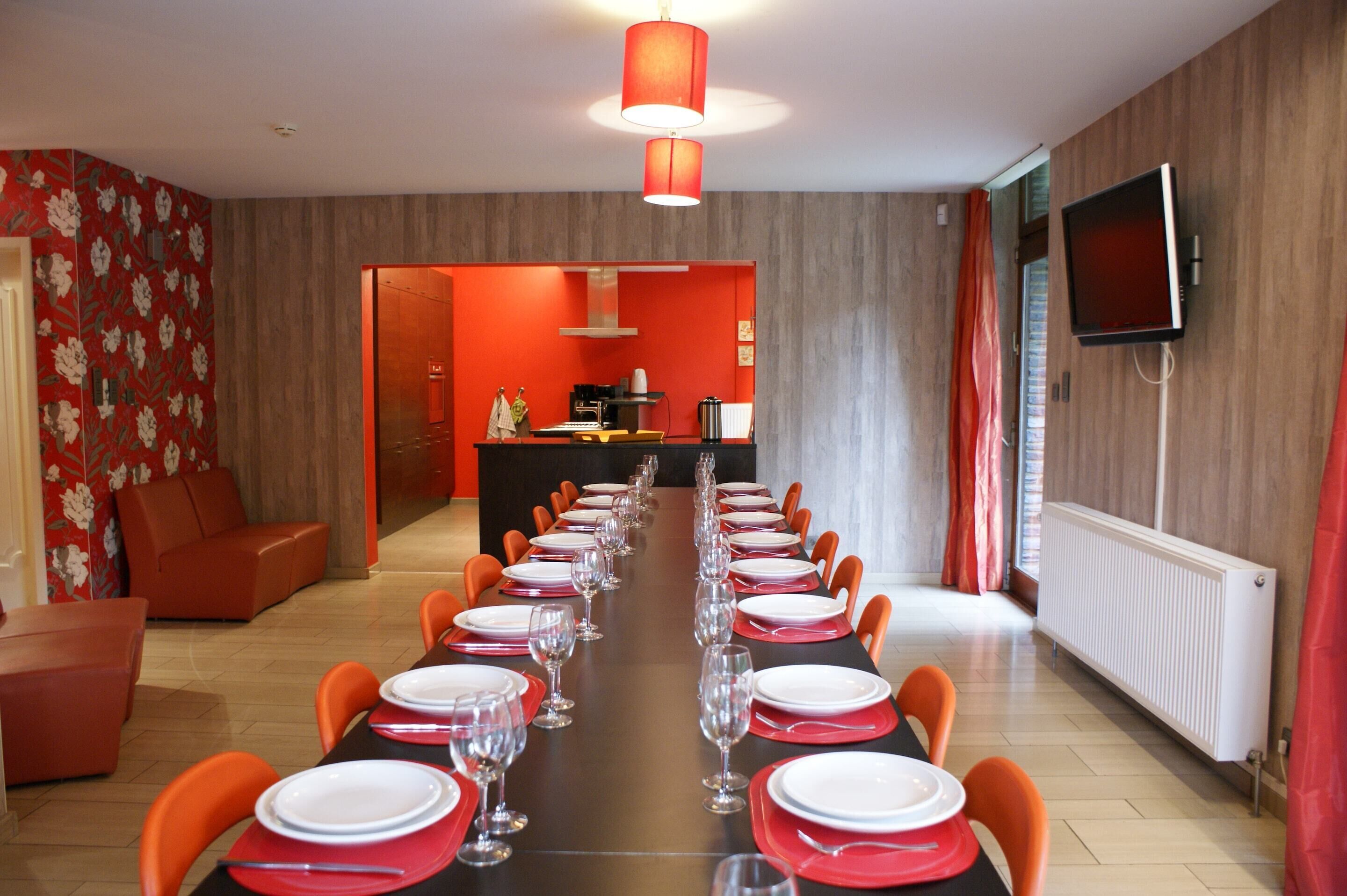 A dining area with a long table set for a meal, a kitchen in the background, and a TV mounted on the wall.