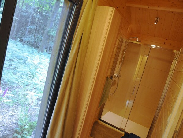 Una cabaña de madera con mampara de ducha de vidrio, una ventana con cortinas y vistas a un área boscosa.