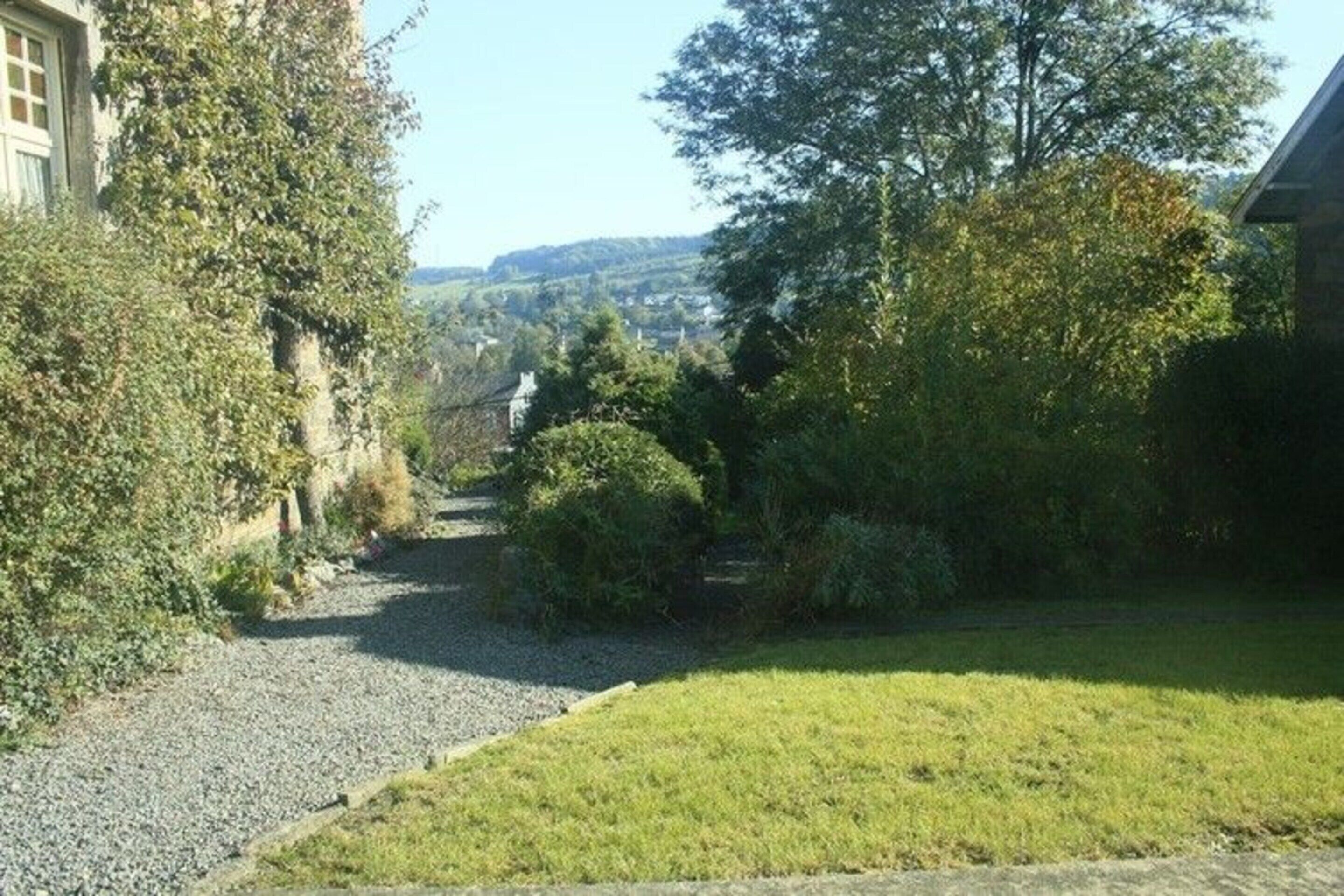 Un chemin de gravier traverse un jardin à la végétation luxuriante, avec un bâtiment situé sur la gauche.